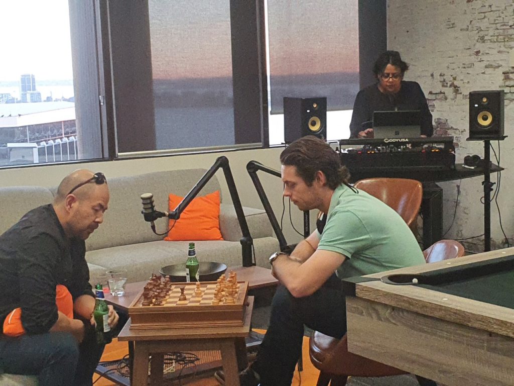 casual pic of 2 men playing chess in the foreground, a women DJ-ing behind them and pool table visible in the corner.